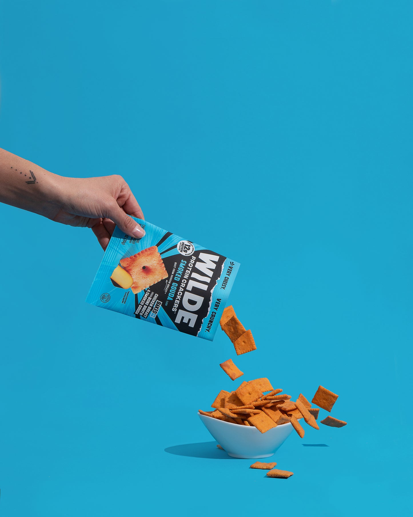 Angled view of Wilde Smoked Gouda protein crackers bag pouring orange crackers into a white bowl against a bright blue background