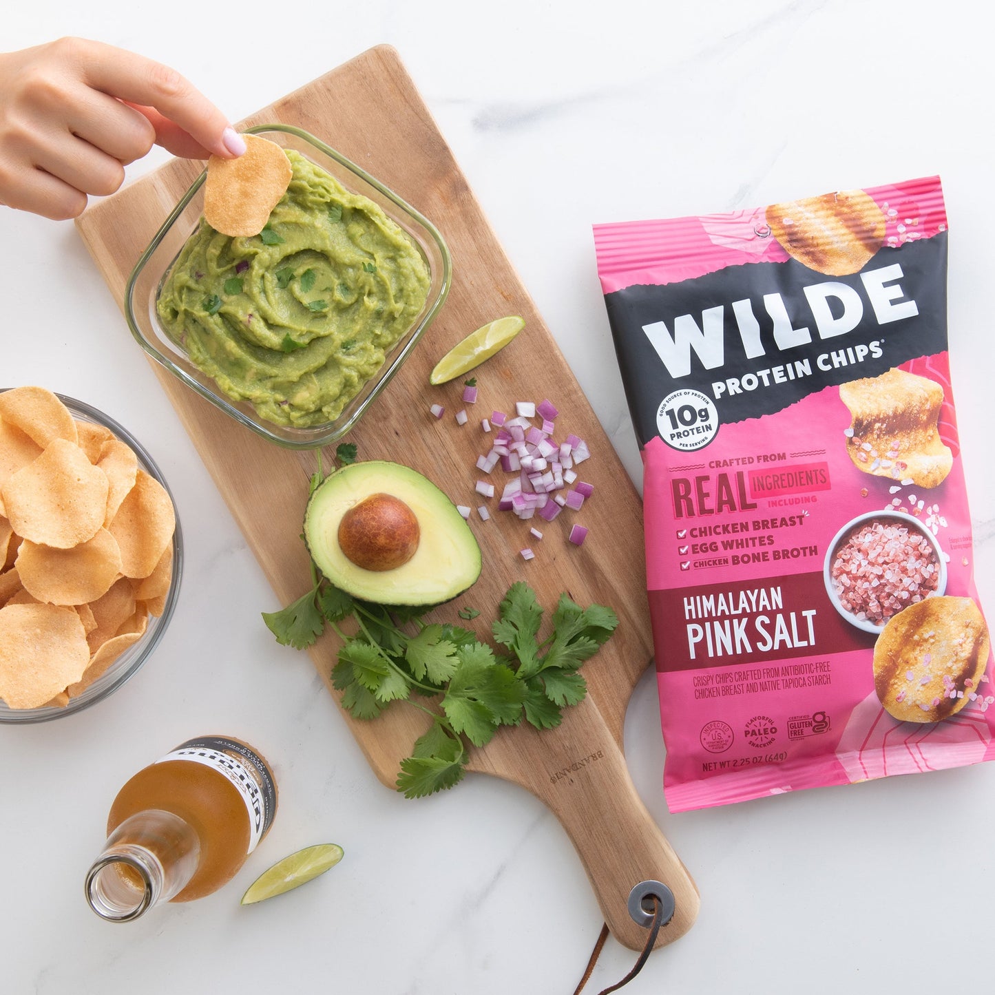 A bowl of guacamole with a chip, avocado, red onion, and Wilde Protein Chips bag labeled 'Himalayan Pink Salt'.