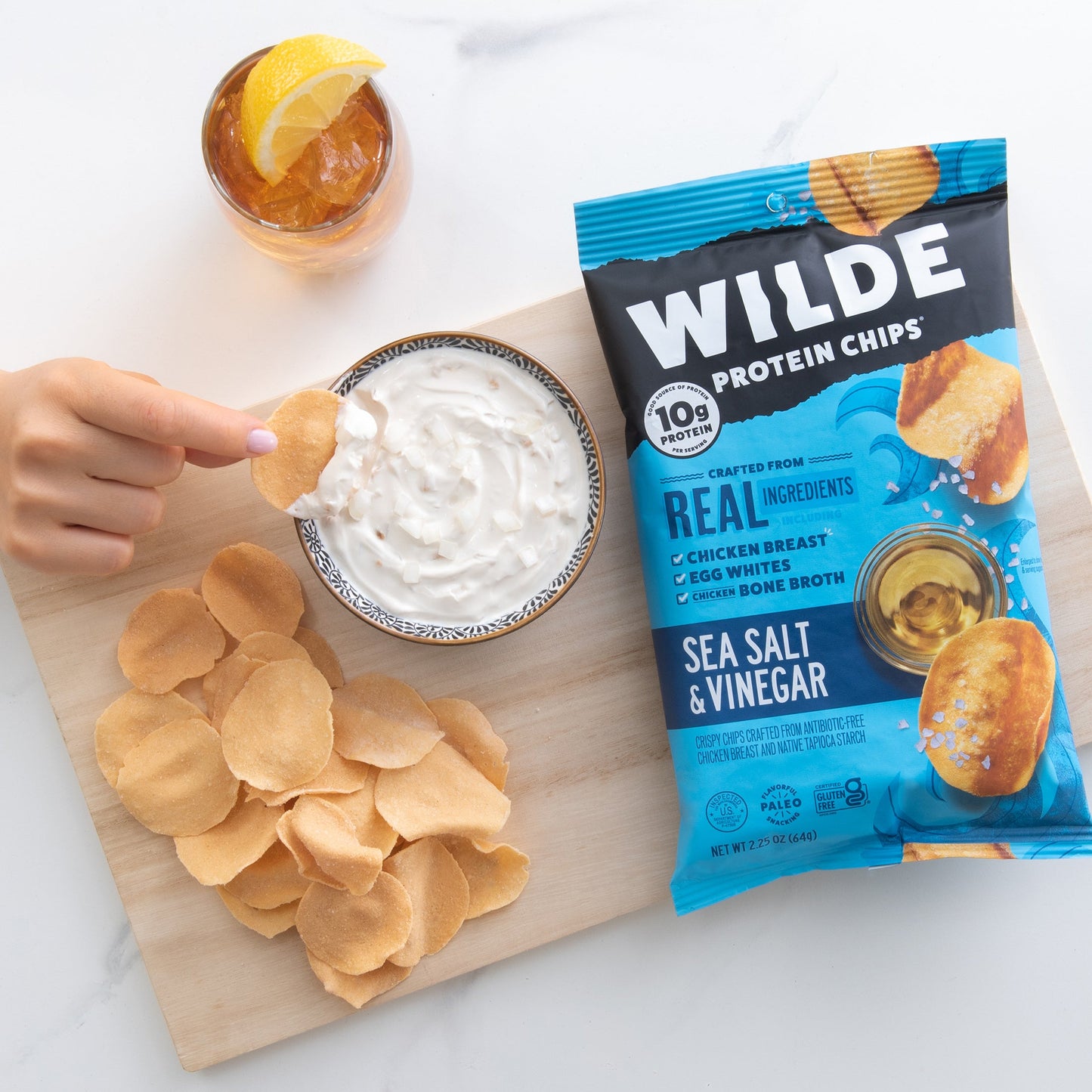 Wilde Protein Chips Sea Salt & Vinegar flavor bag with chips on a wooden board, a hand dipping a chip into a bowl of creamy dip, and a glass of iced tea with a lemon wedge.