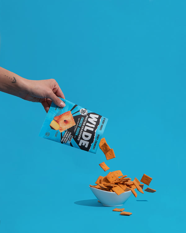 Angled view of Wilde Smoked Gouda protein crackers bag pouring orange crackers into a white bowl against a bright blue background