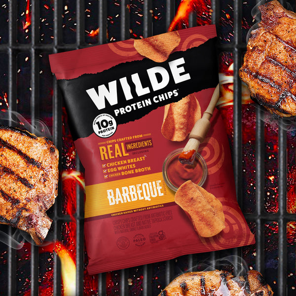 Wilde Protein Chips Barbeque flavor bag on a grill with grilled meat pieces and a brush with barbeque sauce