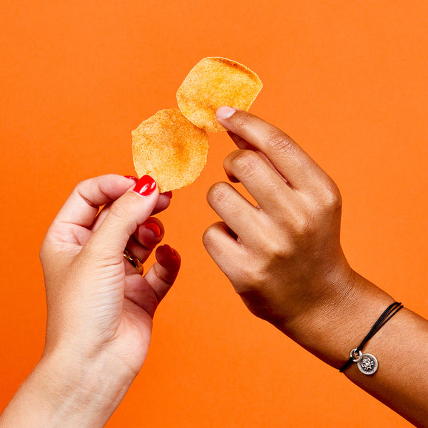 Two hands holding two seasoned potato chips against an orange background, one hand with red nail polish and the other wearing a black string bracelet with a silver charm
