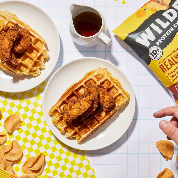 Waffle with fried chicken on plate, syrup pitcher, chips, and hand by bag labeled WILD PROTEIN CHIPS REAL CHICKEN 10g